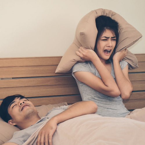 Woman using a pillow to block out noise from a snoring man in bed.
